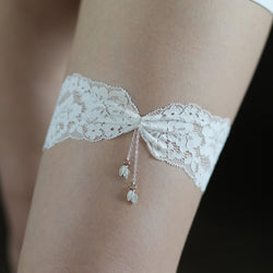 Close-up on-body view of the intricate white floral lace, central twisted knot, and shimmering chain fringe tassels on the Arachne Elysian bridal garter.