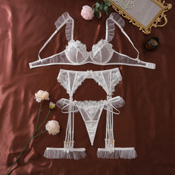 Flat lay presentation of the complete KissKismet white floral embroidered lingerie set, including bra, thong, and garter belt on a satin background.